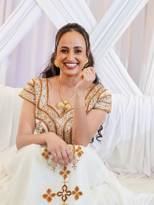 Woman wearing a white and gold embroidered traditional Habesha Kemis with a decorative background.