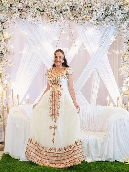 Woman in a white and gold embroidered Habesha Kemis standing in front of a decorated floral arch.