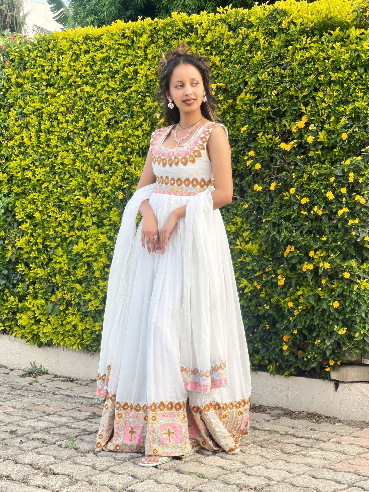 Woman in a traditional Ethiopian Dress standing in front of a green hedge with yellow flowers.