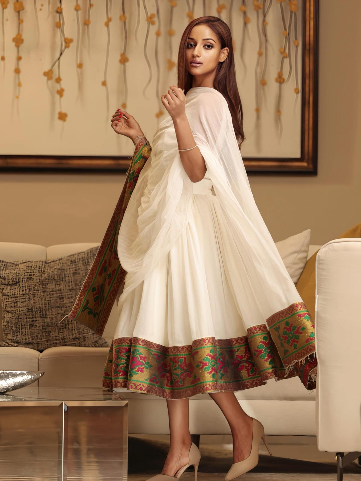 A woman models a white traditional three-piece Ethiopian Dress with a heavily embroidered multicolor border on the hem and dupatta, set in a warm, modern living room.