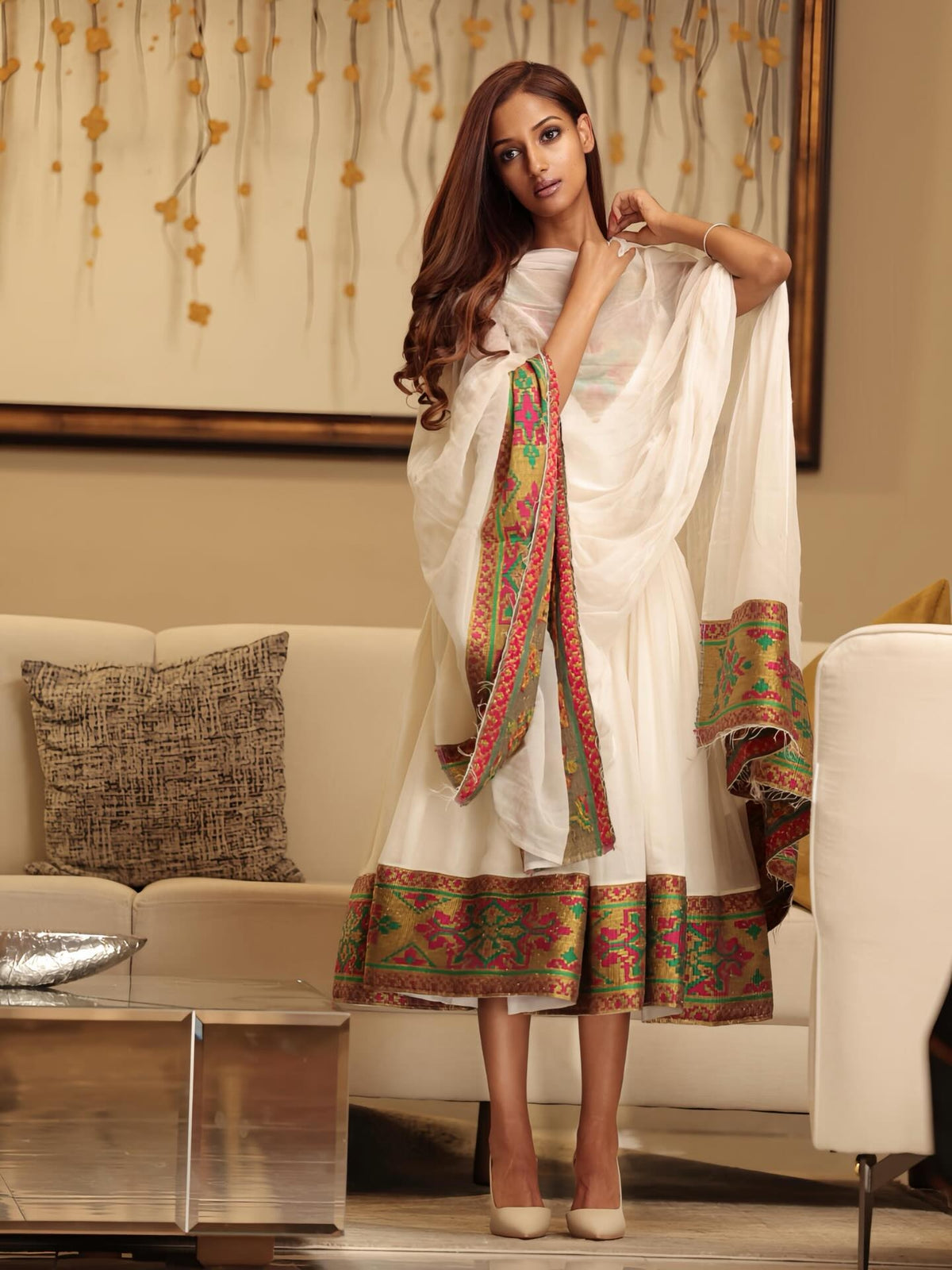 A woman models a white traditional Ethiopian Dress with a matching dupatta, featuring ornate multicolor embroidered borders in red, green, and gold. She is standing in a modern living room setting with a beige sofa and a decorative wall art piece in the background.