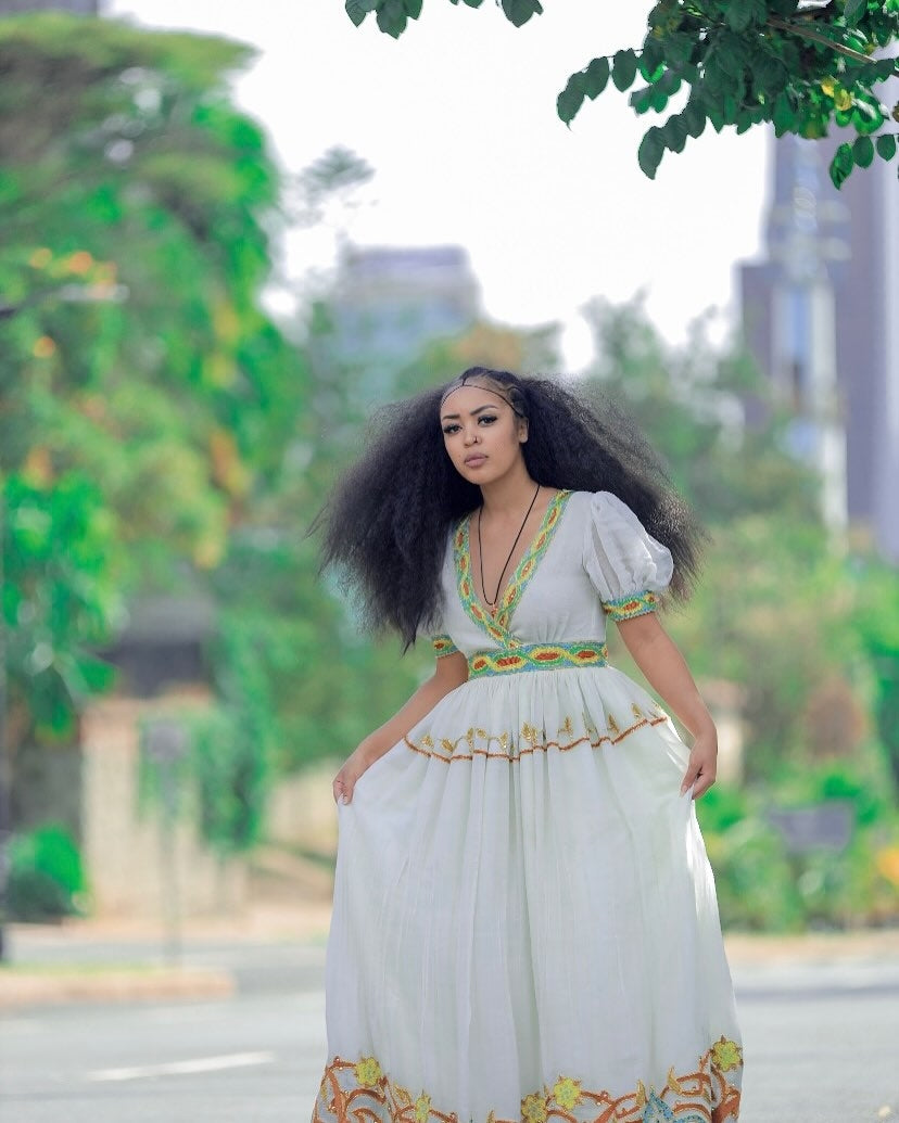 Woman in a white dress with colorful embroidery standing outdoors with greenery in the background
