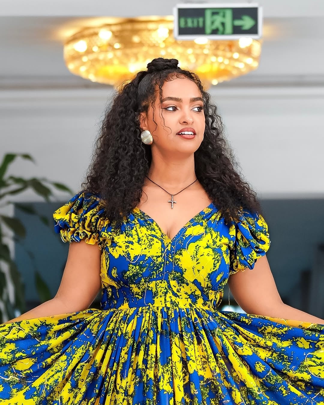 Woman wearing a blue and yellow patterned Yellow and Green Shiffon Ethiopian Dress indoors with a chandelier in the background.