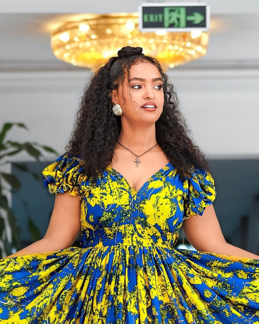 Woman wearing a blue and yellow patterned Yellow and Green Shiffon Ethiopian Dress indoors with a chandelier in the background.