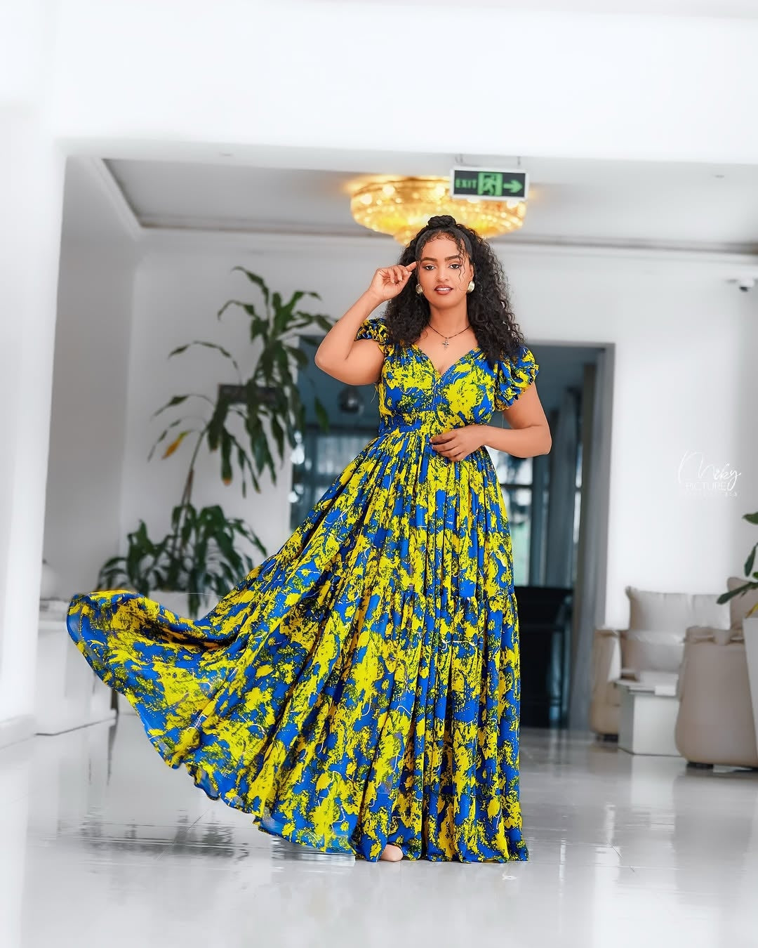 Woman in a blue and yellow floral Yellow and Green Shiffon Ethiopian Dress standing in a modern indoor setting.