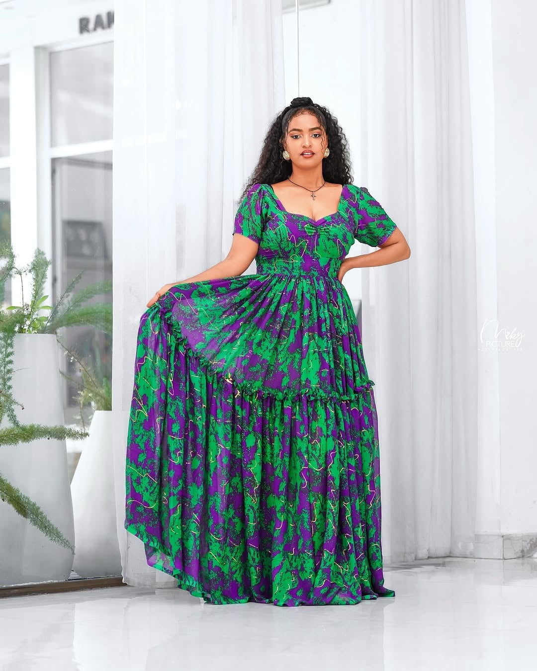 Woman wearing a green and purple Green Habesha Shifion  in a bright room with white curtains.