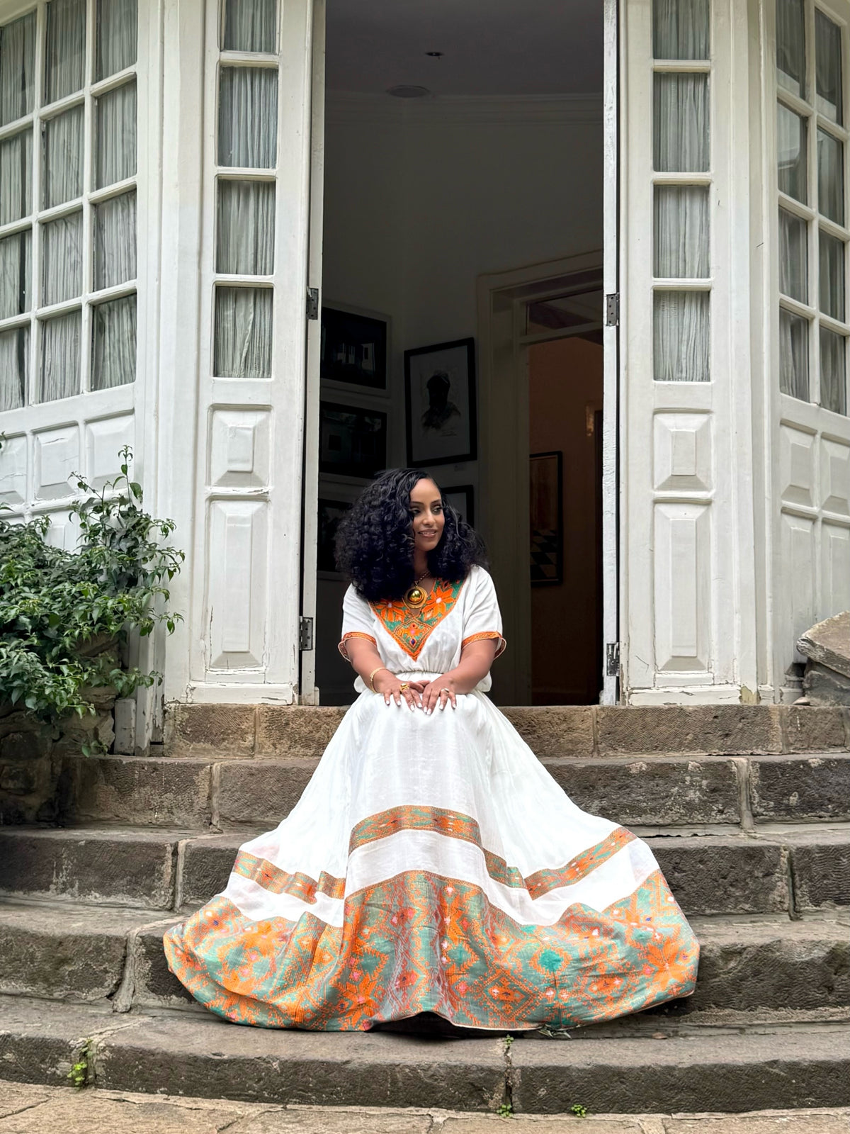 Woman in a white Ethiopian Dress with colorful patterns sitting on steps outside a building.