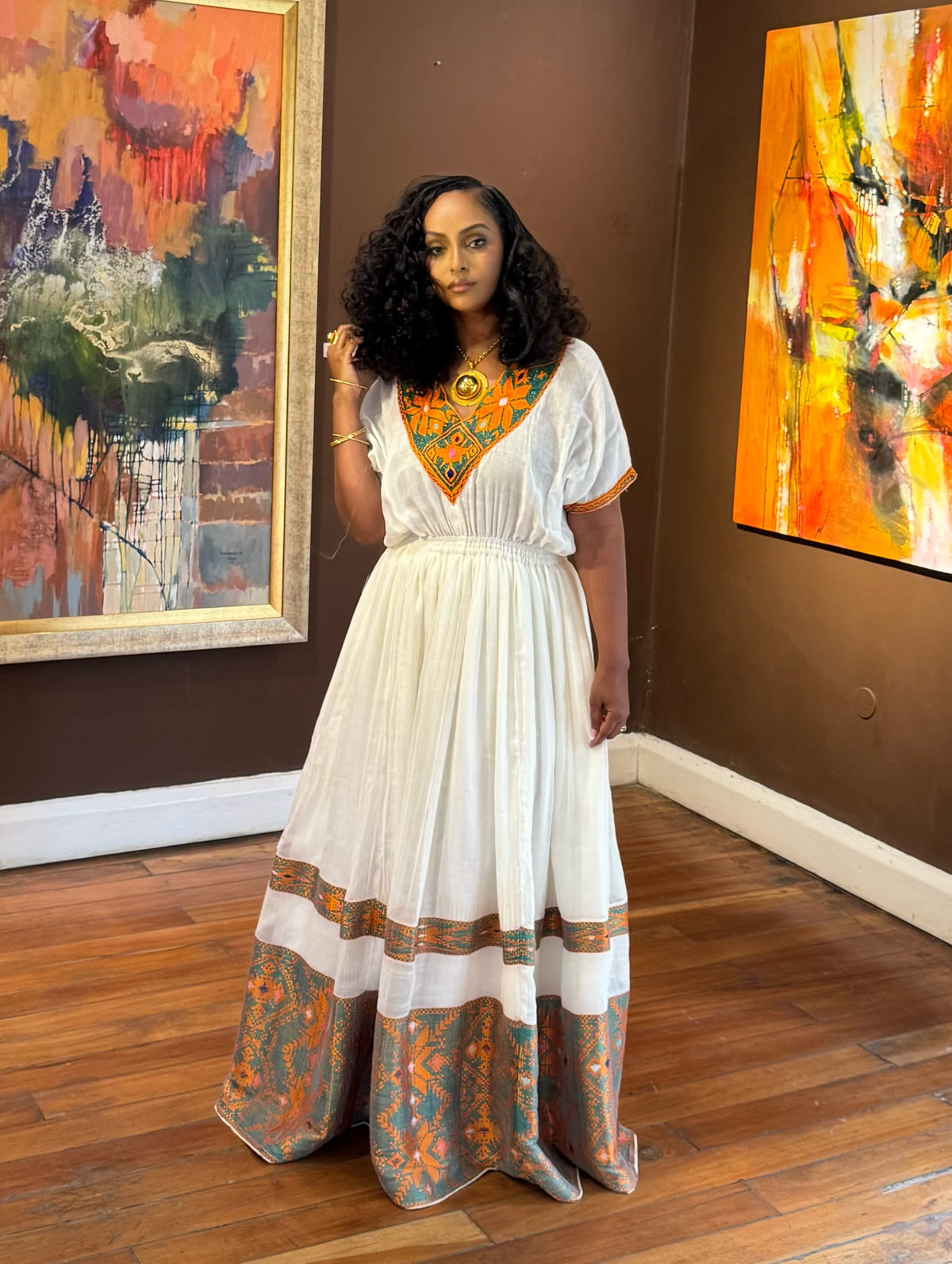 Woman in a white Ethiopian Dress with orange and blue patterns standing in front of abstract paintings.
