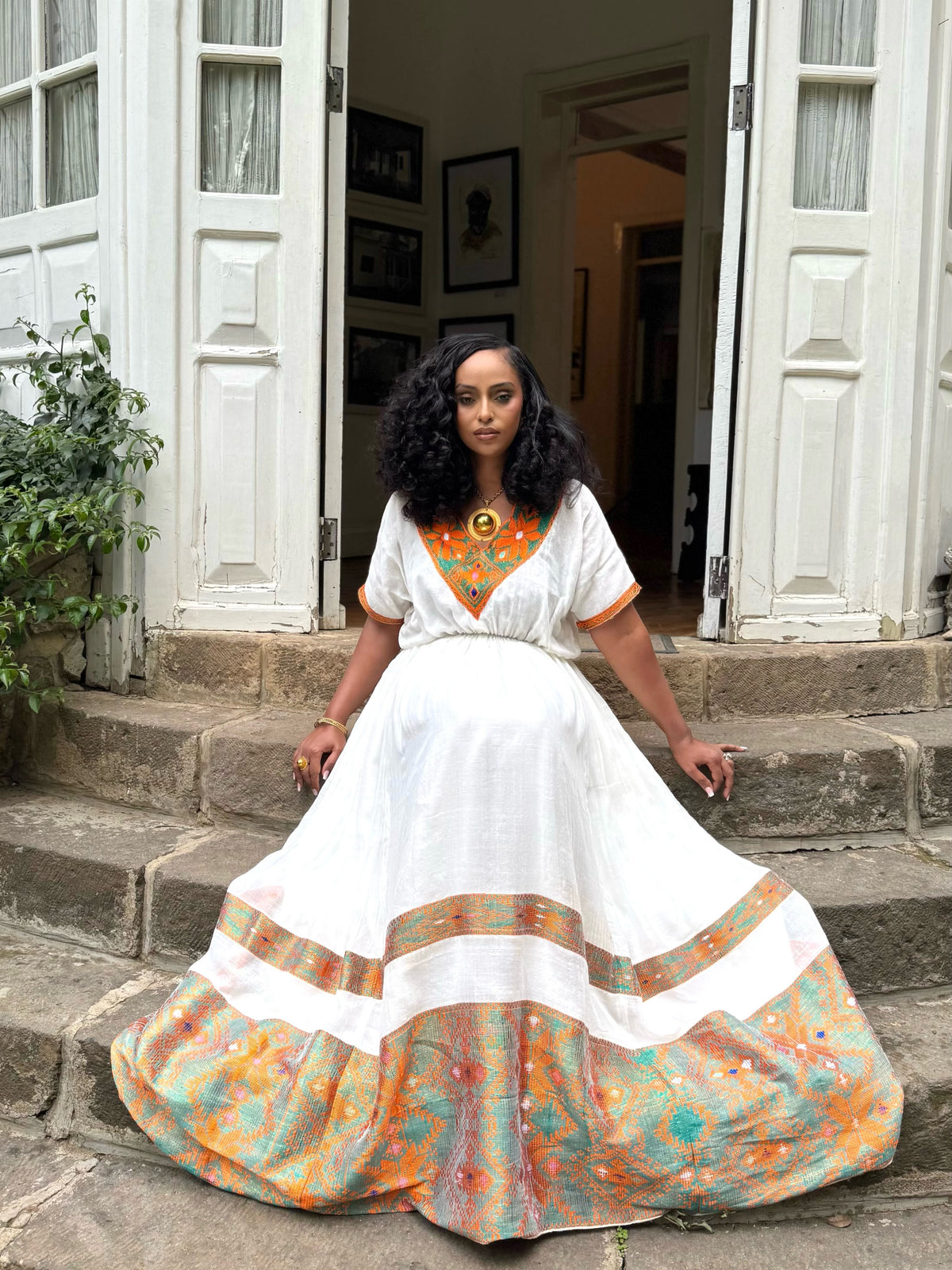 Woman in a white Ethiopian Dress with colorful patterns sitting on steps outside a house.
