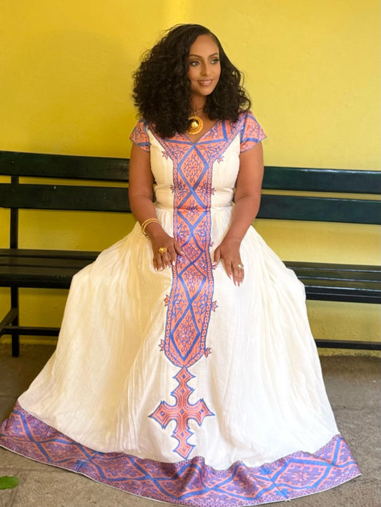 A woman wearing a traditional white Afar habesha zuria with blue and pink geometric patterns, sitting on a dark green bench against a yellow wall.