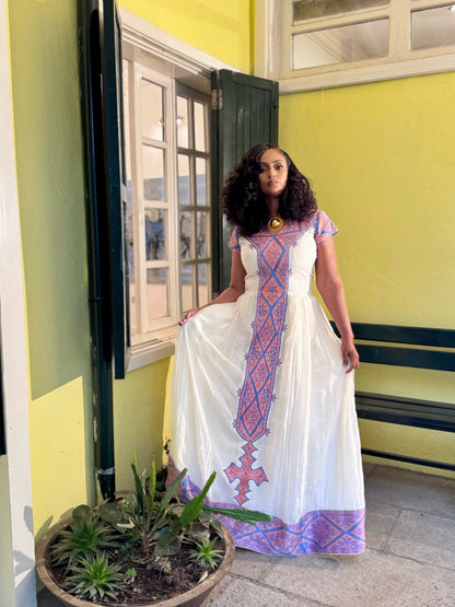 A woman wearing a white habesha zuria  with pink and purple geometric embroidery, standing in front of a yellow wall with a window and bench.