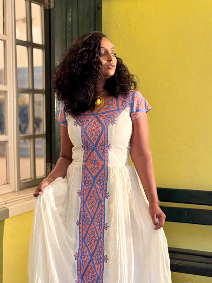 A woman wearing a white habesha zuria  with blue and orange geometric embroidery, standing in front of a yellow wall.