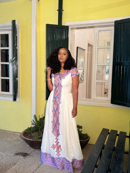 A woman wearing a white habesha zuria  with colorful geometric patterns and a gold necklace, standing in front of a yellow building with dark green shutters.