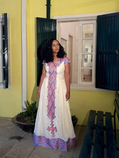 A woman wearing a long white habesha zuria with pink and blue geometric embroidery, standing in front of a yellow wall with dark green shutters.