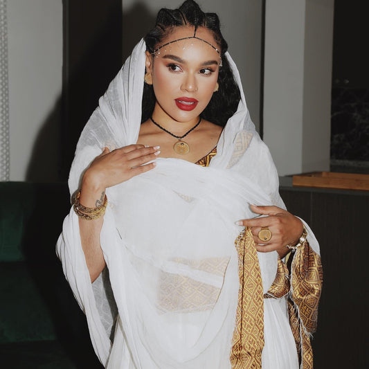 A woman wearing a white draped shawl and a gold patterned Ethiopian Traditional Clothing, styled with gold jewelry including a headpiece, necklace, rings, and bracelets.