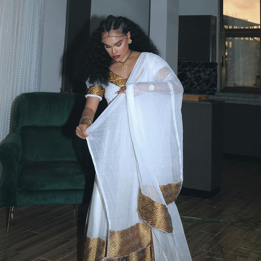 A woman wearing a white  Ethiopian Traditional Clothing with gold patterned trim and a sheer white dupatta, standing in an indoor room with a dark green armchair and window in the background.