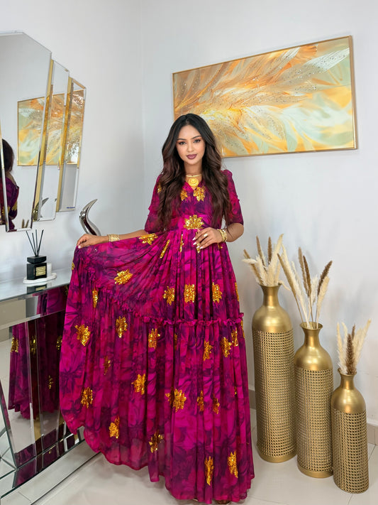 A woman models a long magenta tiered maxi Habesha Chiffon with gold floral patterns in an indoor setting with mirrors, gold vases, and a painting on the wall.