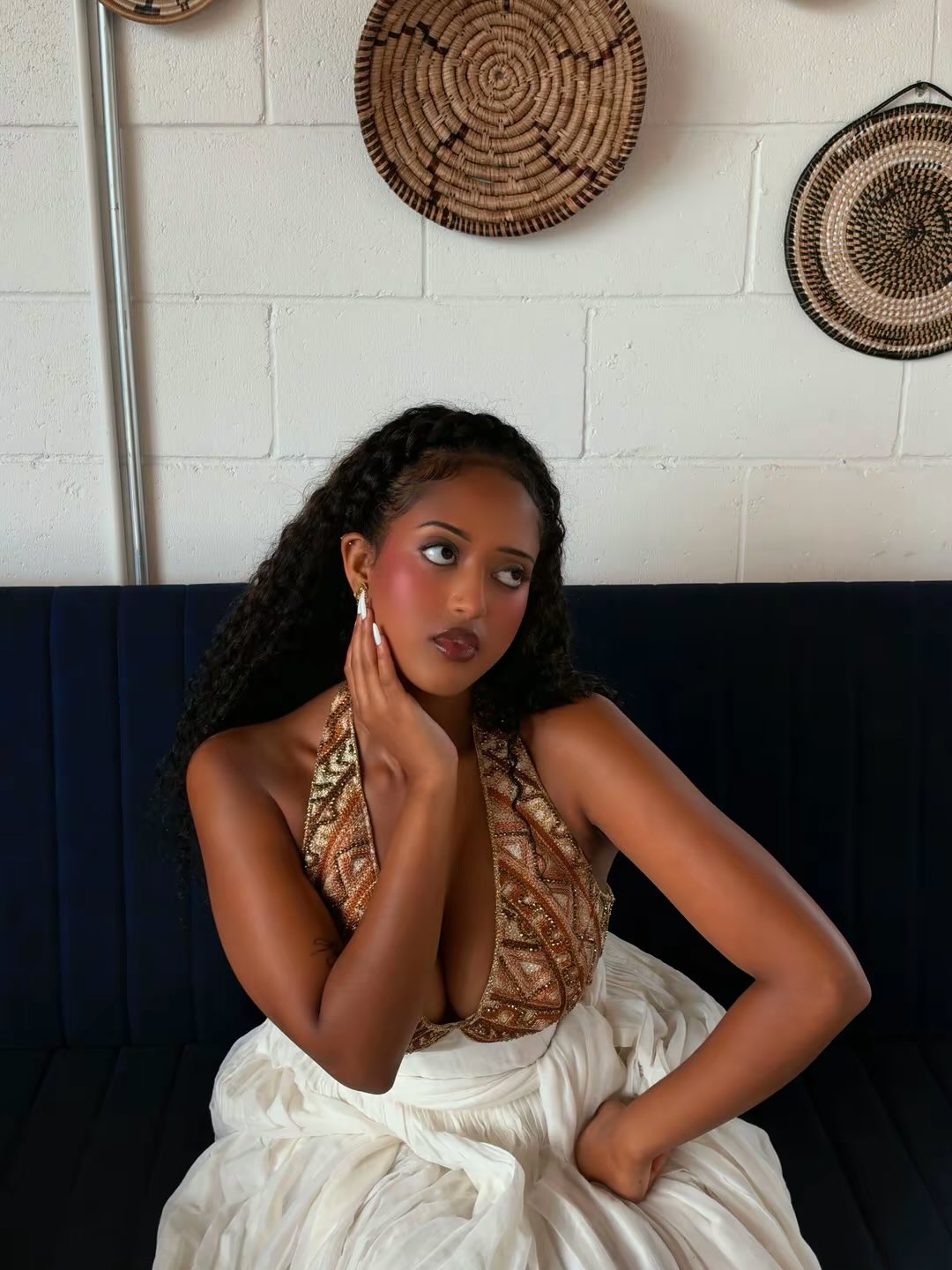 A woman with long dark curly hair sits on a dark navy blue velvet sofa, wearing a sleeveless halter Ethiopian traditional clothing with a brown and gold geometric pattern and a white skirt. She is posed against a white brick wall decorated with woven baskets.