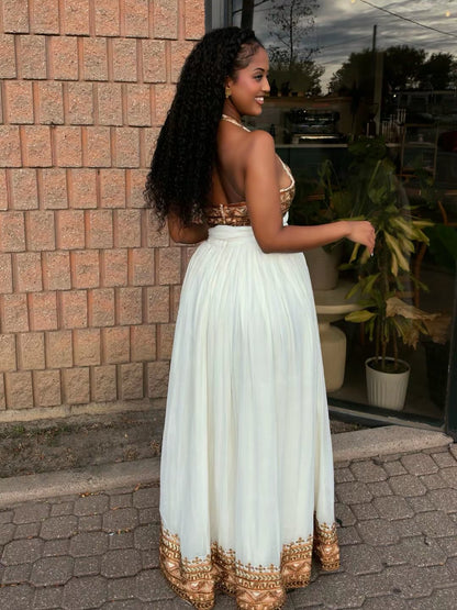 A woman wearing a strapless white Ethiopian traditional clothing with gold embroidery on the bodice and hem, standing outdoors in front of a brick wall and glass door.