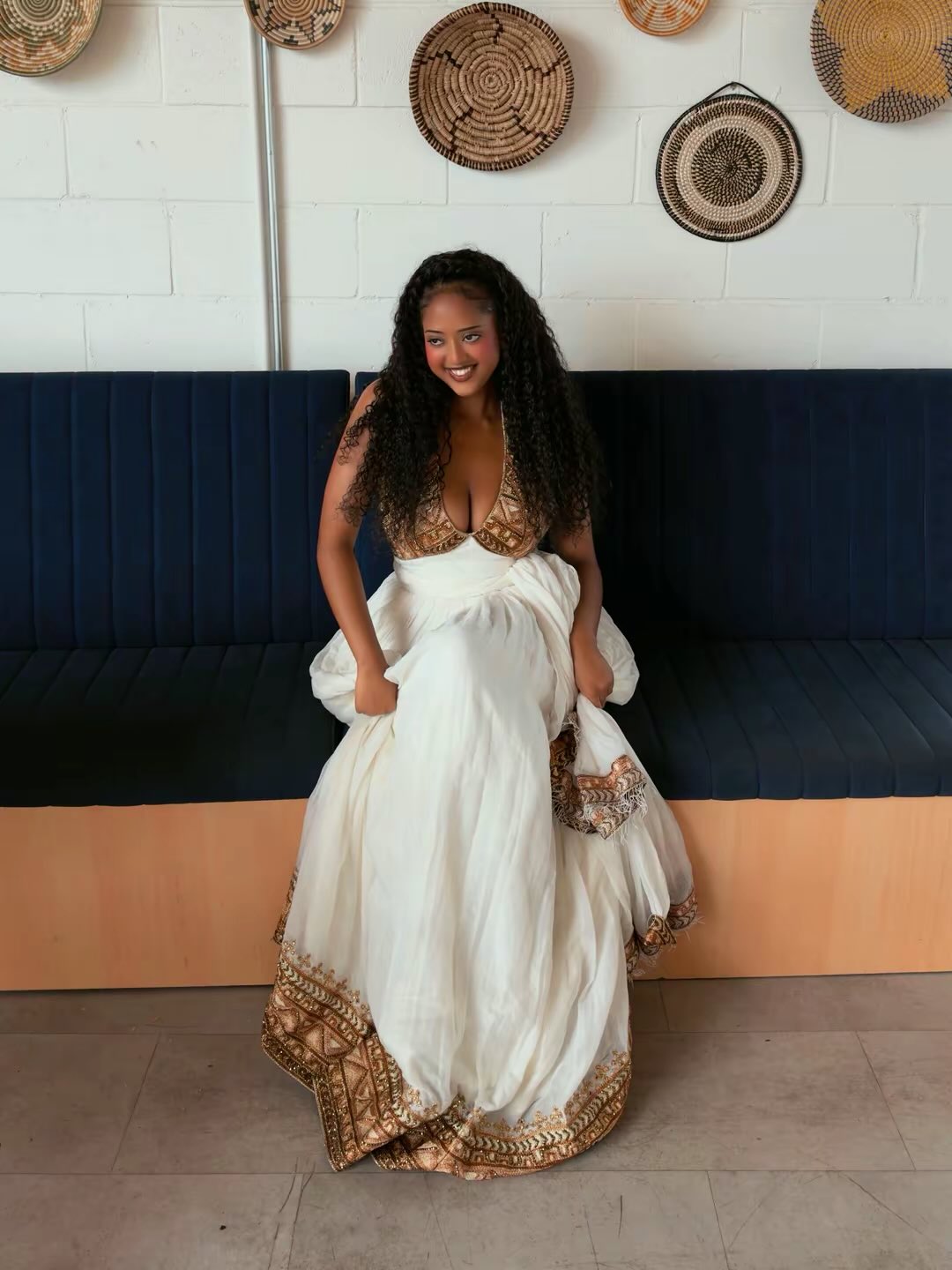 A woman wearing a white Ethiopian traditional clothing with gold embroidery sits on a dark blue bench in front of a white brick wall decorated with woven baskets.