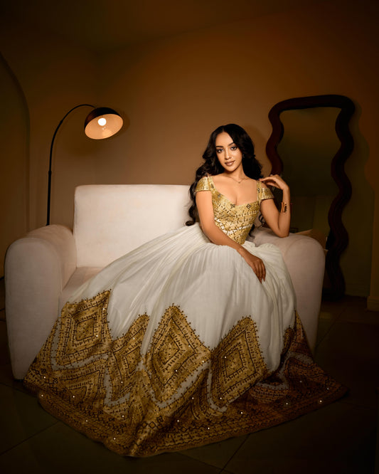 A woman wearing an ornate white and gold traditional Ethiopian traditional clothing sits on a white armchair in a warmly lit room. The dress features a heavily embellished gold bodice and a wide, heavily embroidered gold and white border at the hem.