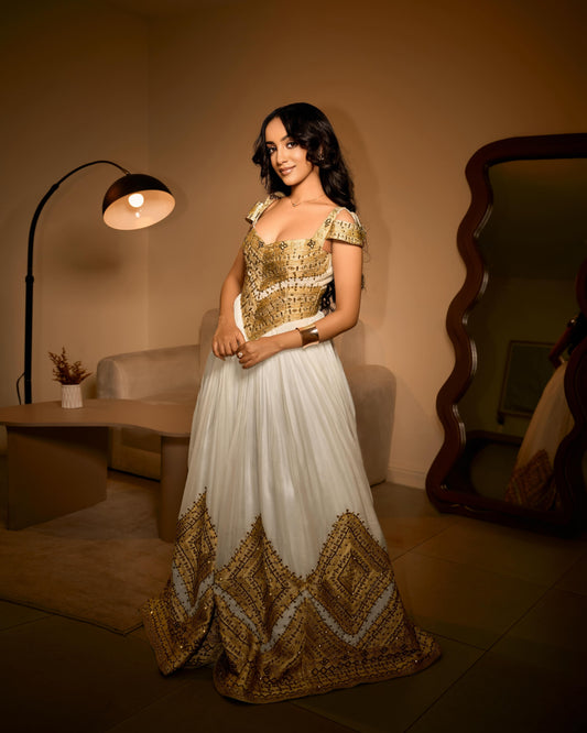 A woman stands in a warmly lit room wearing a formal off-the-shoulder Ethiopian traditional clothing with a gold embellished bodice and a long white skirt featuring a large gold geometric border at the hem.