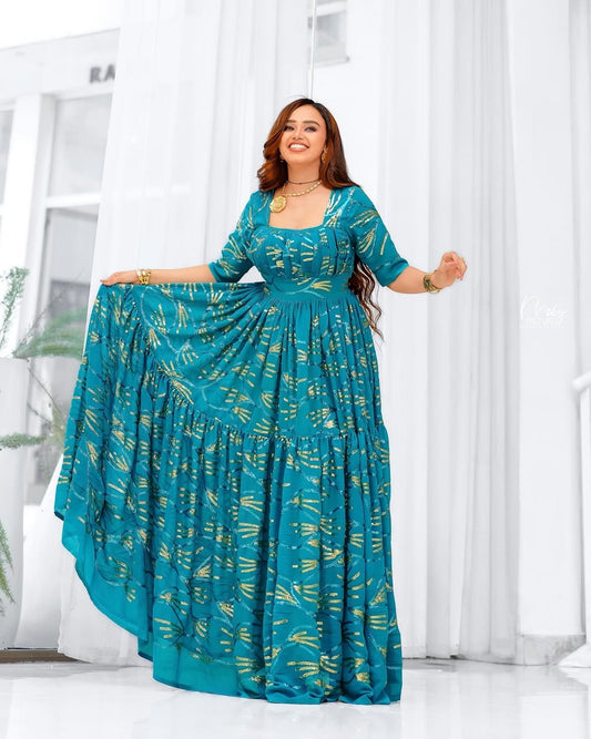 Woman wearing a teal chiffion dress with gold patterns in a bright room.