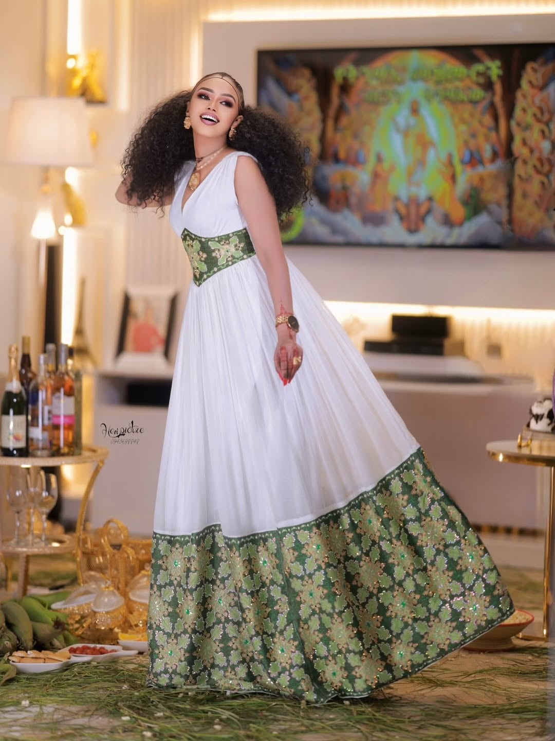 Woman in a white and green Ethiopian dress standing in a decorated room with a colorful painting on the wall.