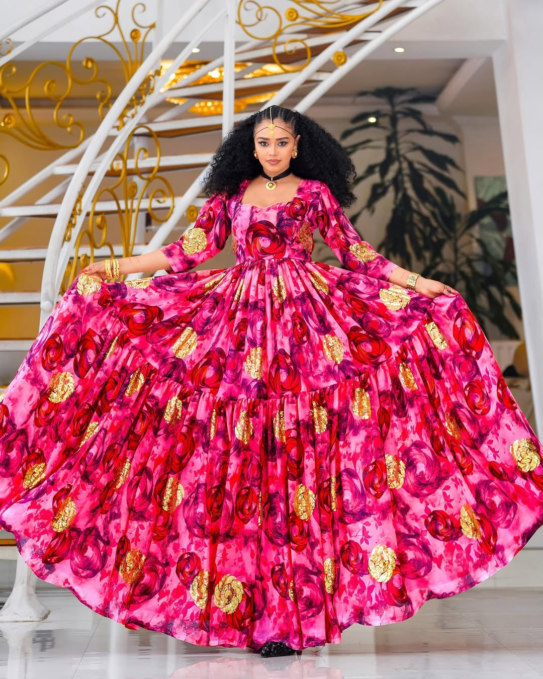 Woman in a vibrant pink Habesha Chiffon with red floral patterns and gold accents standing in a decorative interior setting.