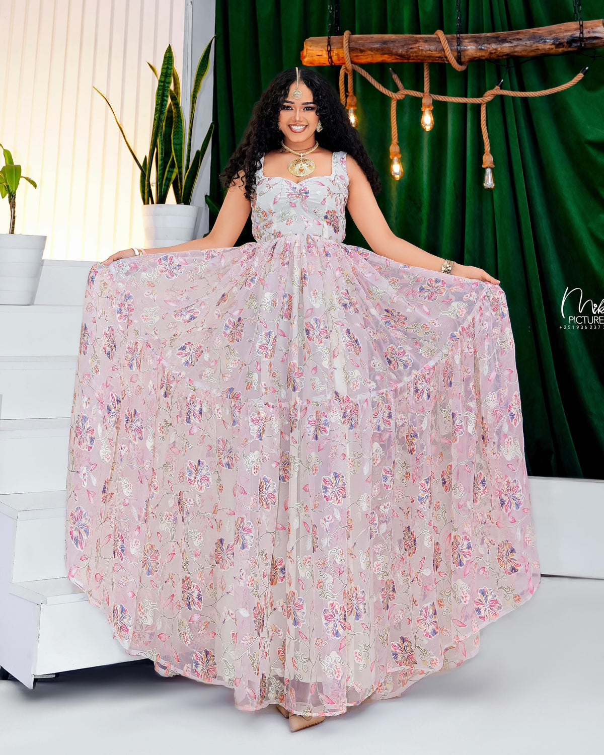 Woman in a floral Habesha Kemis posing in front of a green curtain with decorative lights.