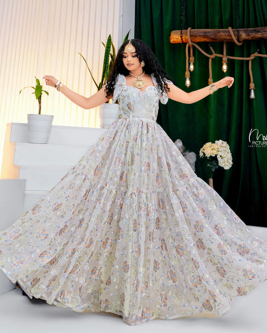 A woman modeling a voluminous off-white evening chiffon Habesha with a delicate floral pattern and sheer off-the-shoulder straps, posed against a backdrop of a green curtain and white steps.