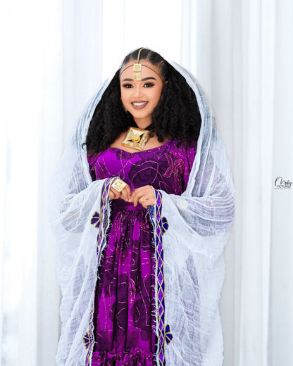 A woman wearing a traditional purple  Habesha chiffon  with gold embroidery, a white sheer veil, and gold jewelry, standing in front of a white curtain background.