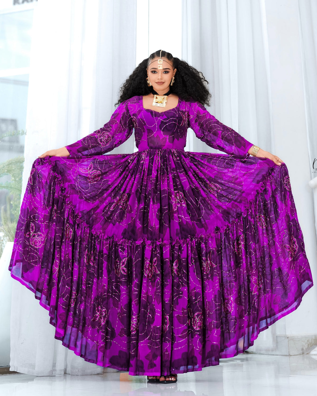 A woman modeling a vibrant purple long-sleeve  Habesha chiffon  with a tiered ruffled skirt, holding the fabric outwards to show its volume against a bright white curtain background.
