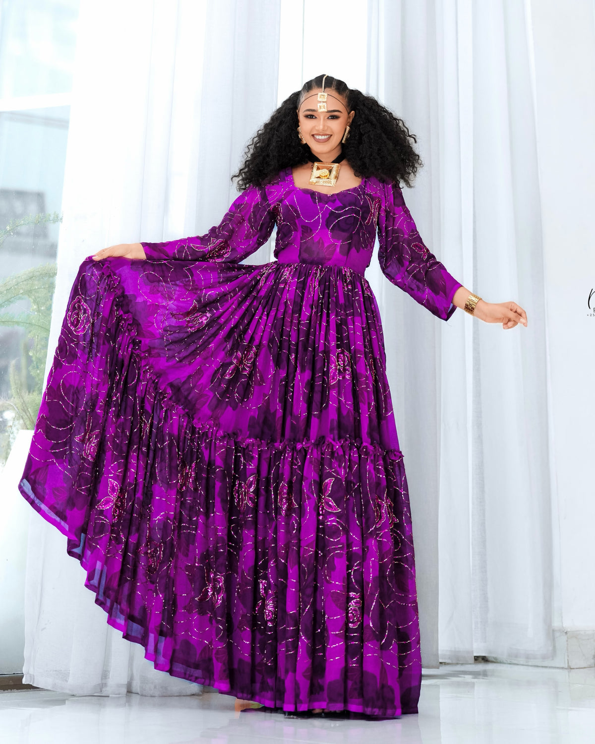 A woman modeling a long-sleeved magenta purple  Habesha chiffon  with a metallic gold abstract pattern, standing in front of white curtains.