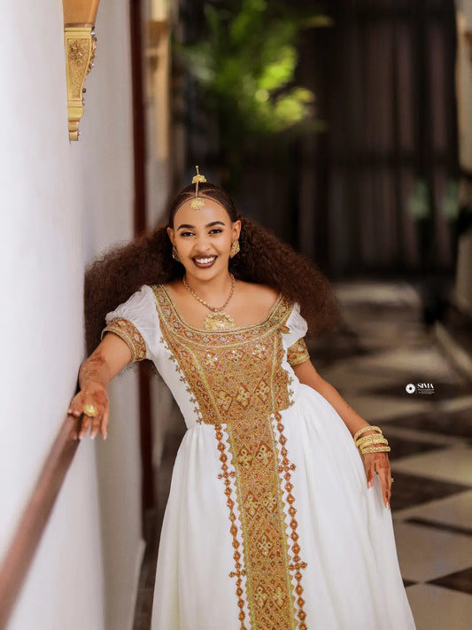 A woman wearing a traditional white Ethiopian dress with ornate gold embroidery and gold jewelry, posing in a corridor with a white wall on the left.