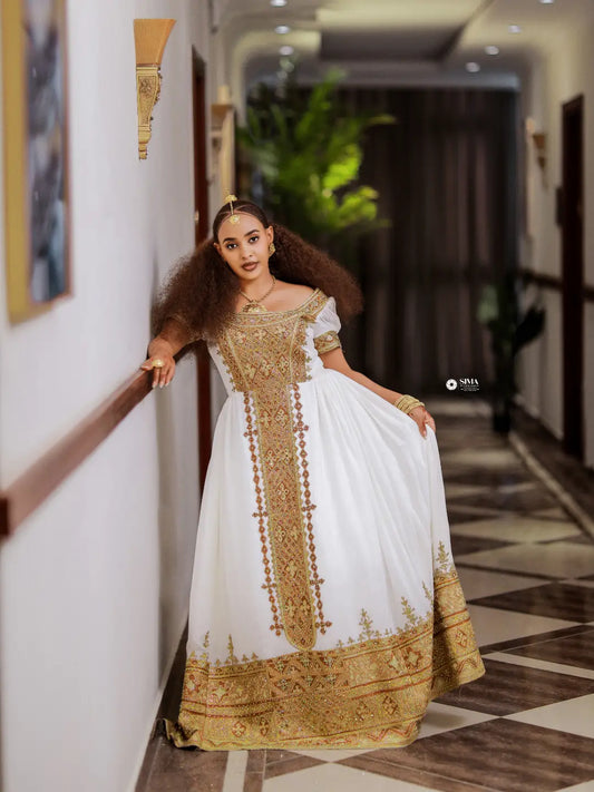 A woman wearing an elegant white and gold embroidered traditional Ethiopian dress stands in a hallway, holding out one side of the gown to show its full width. The dress features intricate gold embroidery on the bodice, front panel, sleeves, and hem.