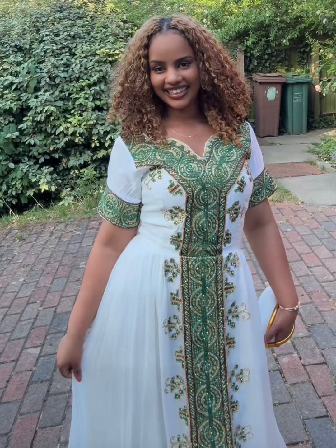 Woman wearing a white habesha dress with green and gold embroidery standing outdoors.