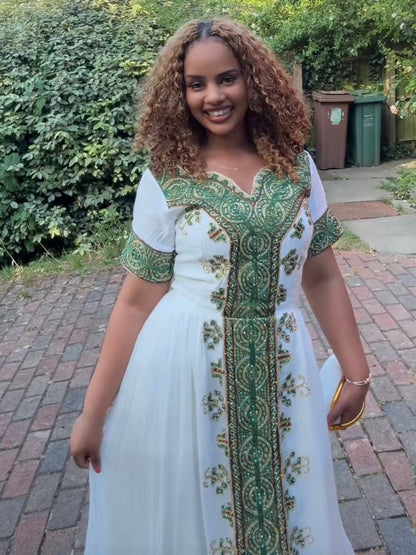 Woman wearing a white habesha dress with green and gold embroidery standing outdoors.