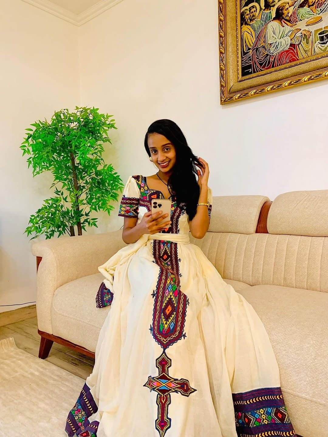 Woman in a colorful habesha dress sitting on a couch in a living room.