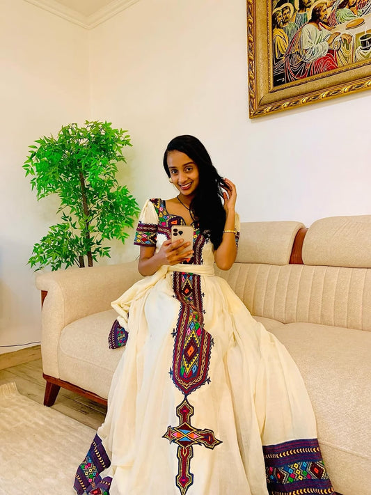 Woman in a colorful habesha dress sitting on a couch in a living room.