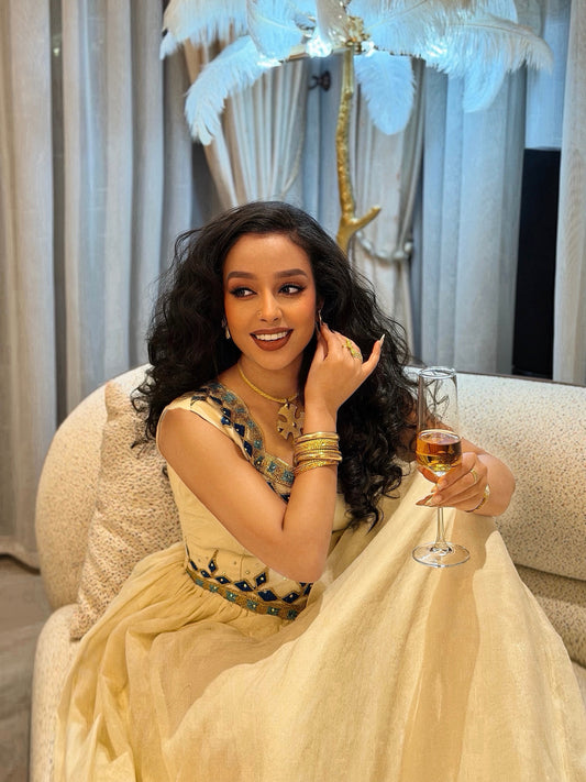 A woman with long, wavy black hair sits on a beige armchair, wearing a cream-colored traditional dress with dark blue and gold embroidery. She is holding a glass of champagne in a decorated indoor setting with curtains and a feather lamp.