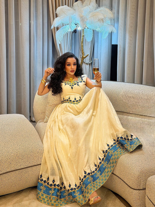 A woman sits on a beige sofa wearing a cream-colored traditional gown with blue and gold patterned borders and a matching dupatta. She holds a glass of champagne in a decorated indoor setting with curtains and a feathered lamp.