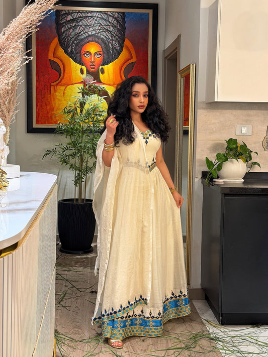 A woman stands in a dimly lit interior wearing an off-the-shoulder formal Woman wearing a traditional embroidered Habesha kemis in a dimly lit room with warm lighting. with a cream tulle skirt, heavy red and gold embroidery, and a heavily embellished dark red and gold flared hem.