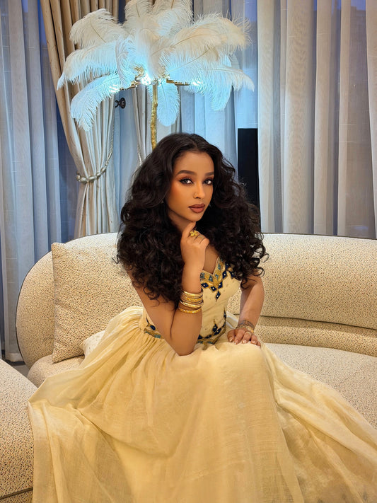 A woman stands in a dimly lit interior wearing an off-the-shoulder formal Woman wearing a traditional embroidered Habesha kemis in a dimly lit room with warm lighting. with a cream tulle skirt, heavy red and gold embroidery, and a heavily embellished dark red and gold flared hem.