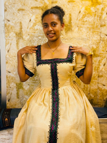 A woman wearing a medieval-style cream-colored Habesha Kemis with black and purple embroidered trim, posing in front of a textured stone wall.