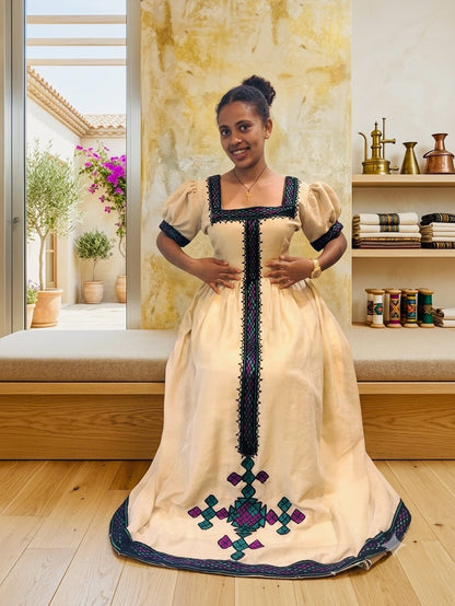 A woman wearing a traditional cream-colored Habesha Kemis with black embroidery and colorful geometric patterns near the hem, standing in a decorated room with wooden floors and shelves.