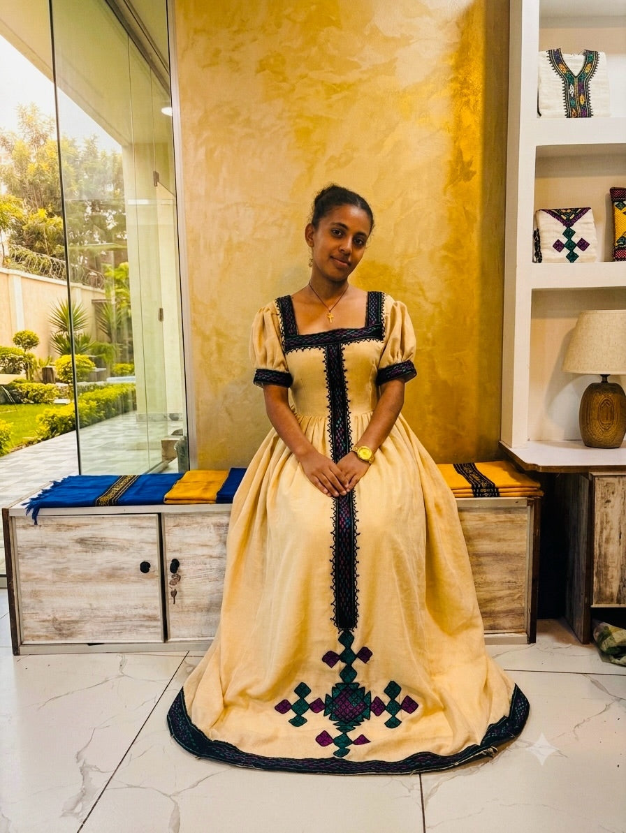 A woman wearing a traditional Ethiopian full-length Habesha Kemis with a beige fabric body, black trim, and colorful geometric embroidery, standing in a decorated room with a gold textured wall.