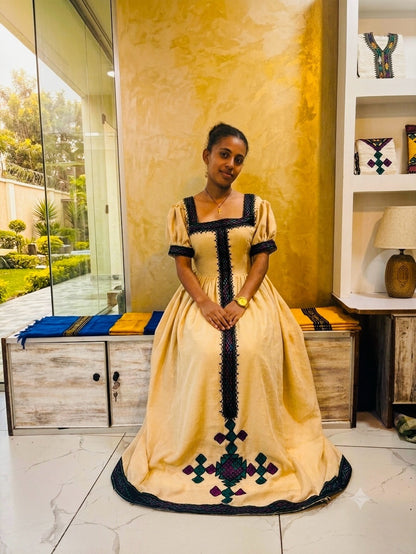 A woman wearing a traditional Ethiopian full-length Habesha Kemis with a beige fabric body, black trim, and colorful geometric embroidery, standing in a decorated room with a gold textured wall.
