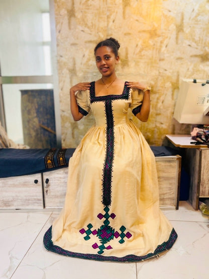 A woman modeling a traditional long Habesha Kemis in a warm cream or pale yellow color with black embroidered trim and a colorful geometric pattern near the hem, standing in an indoor setting.