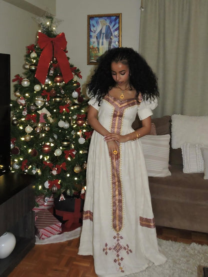 Woman in a long white Habesha Libs
standing in a living room with a decorated Christmas tree.